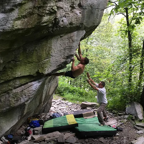 Een shirtloze klimmer is aan het boulderen op een steile rotswand, terwijl een ander persoon beneden staat om hem te begeleiden. Op de grond, omringd door bomen en rotsen, liggen valmatten en klimuitrusting.